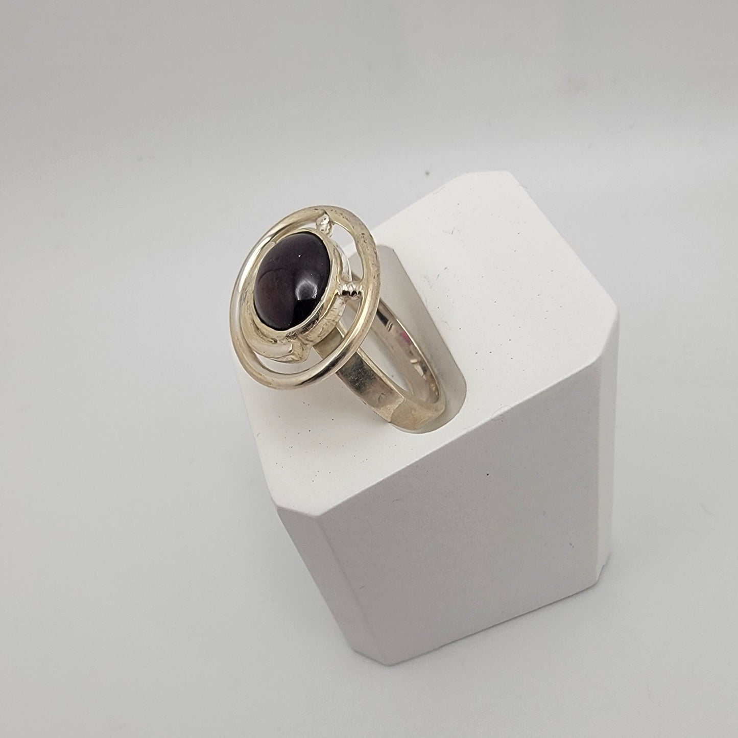 Sterling silver ring with a 12×10 mm oval garnet cabochon set on a uniform 3 mm band, displayed on a white stand.