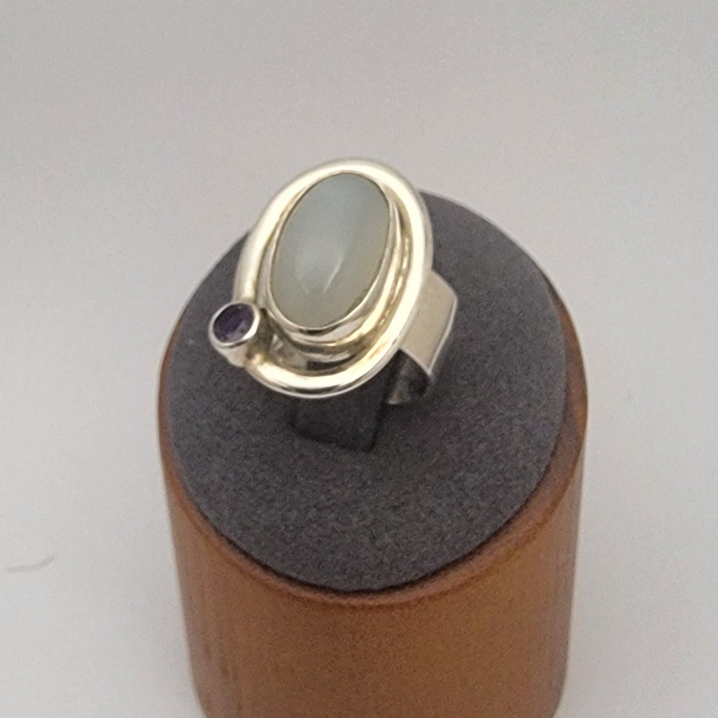 Silver ring with a large moonstone and small amethyst on a gray stand against a white background