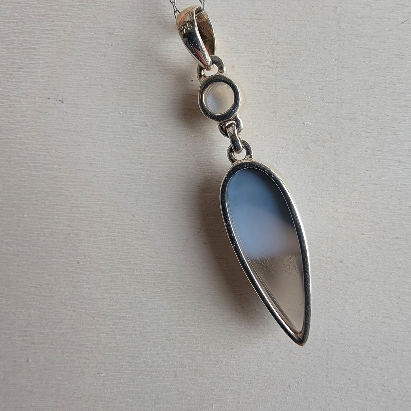 Teardrop-shaped pendant with blue glass and silver chain on a light gray background
