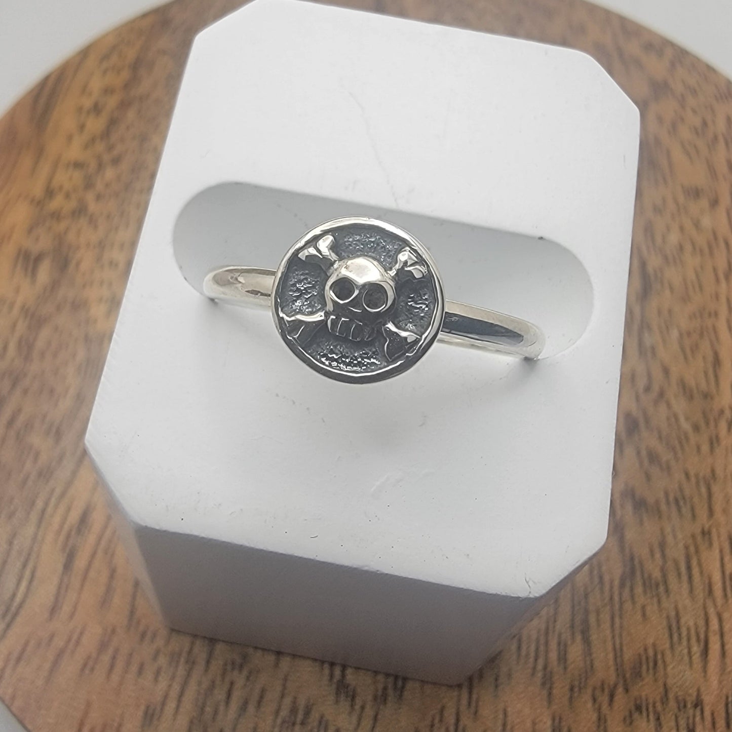 Silver ring with a skull and crossbone design on a white stand against a wooden background