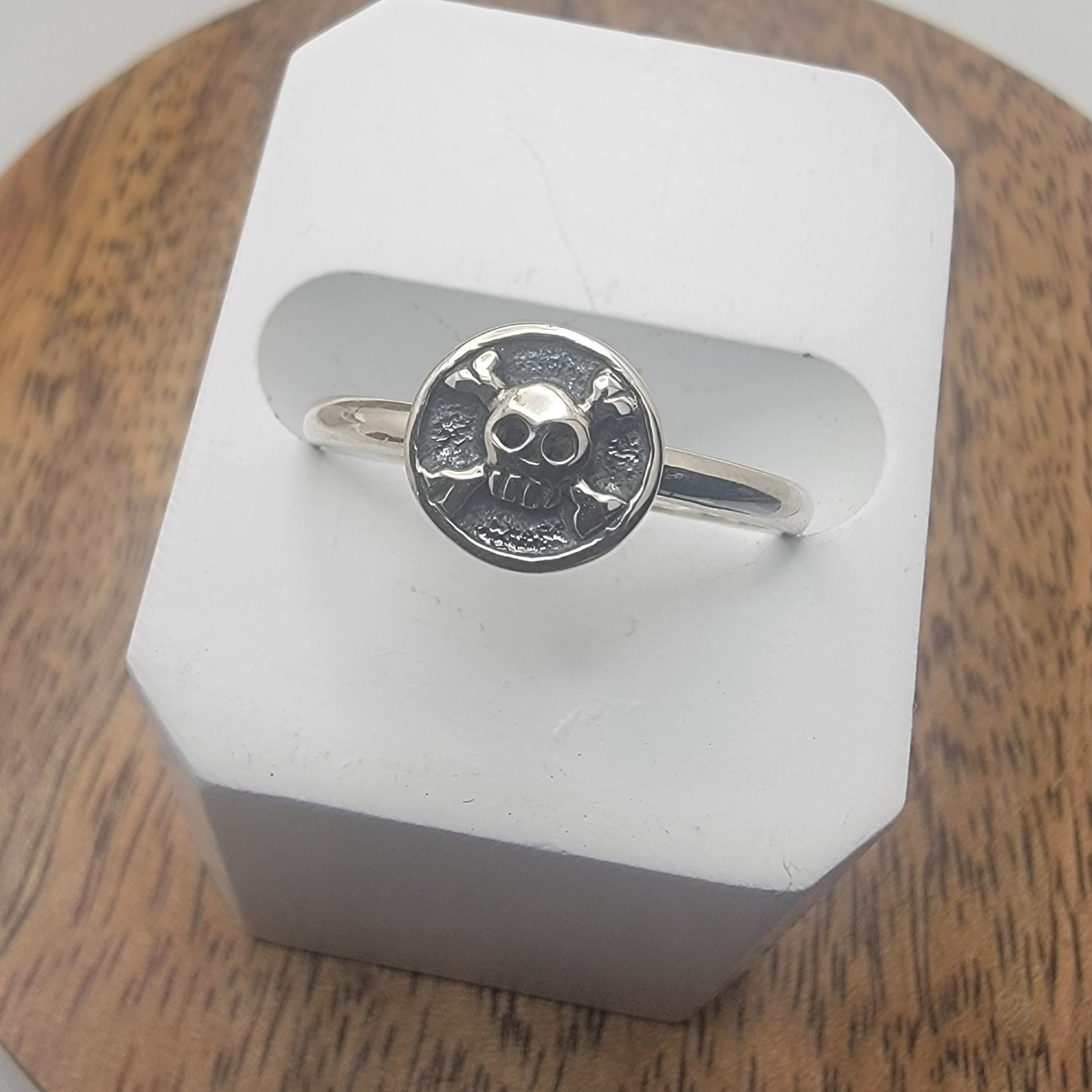 Silver ring with a skull and crossbone design on a white stand against a wooden background