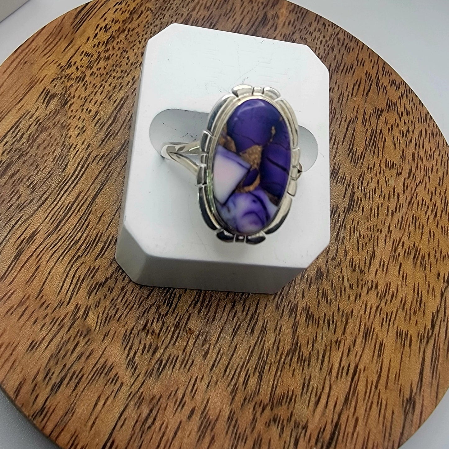 Silver ring with a purple stone on a white stand, placed on a wooden surface.